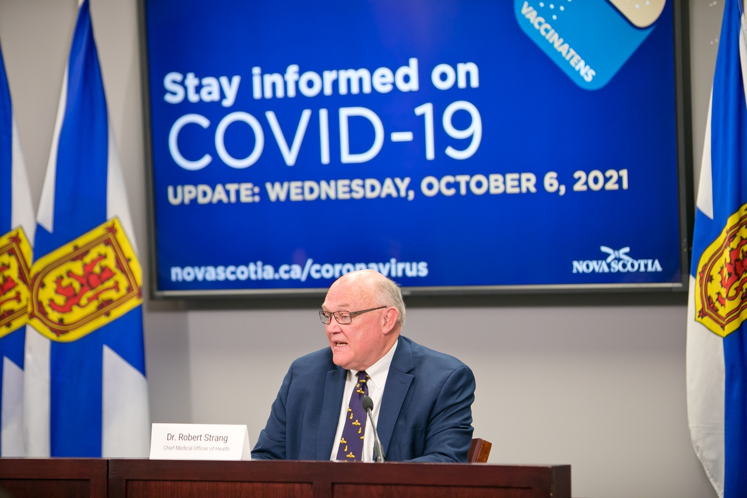 戴眼镜的秃顶男子站在屏幕前，屏幕上写着“随时了解COVID-19”。更新:2021年10月6日，星期三