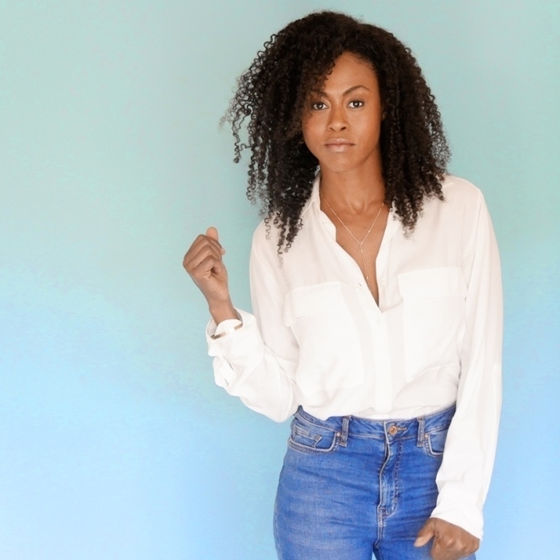 黑人妇女站在一个中性的背景前，穿着牛仔裤和白衬衫