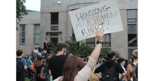 2021年8月18日，在哈利法克斯前纪念图书馆，一群抗议者在警察驱逐无家可归者营地时，一名棕色头发、米白色衬衫的妇女举着一块广告牌，上面用大写字母写着“住房是一项人权!”