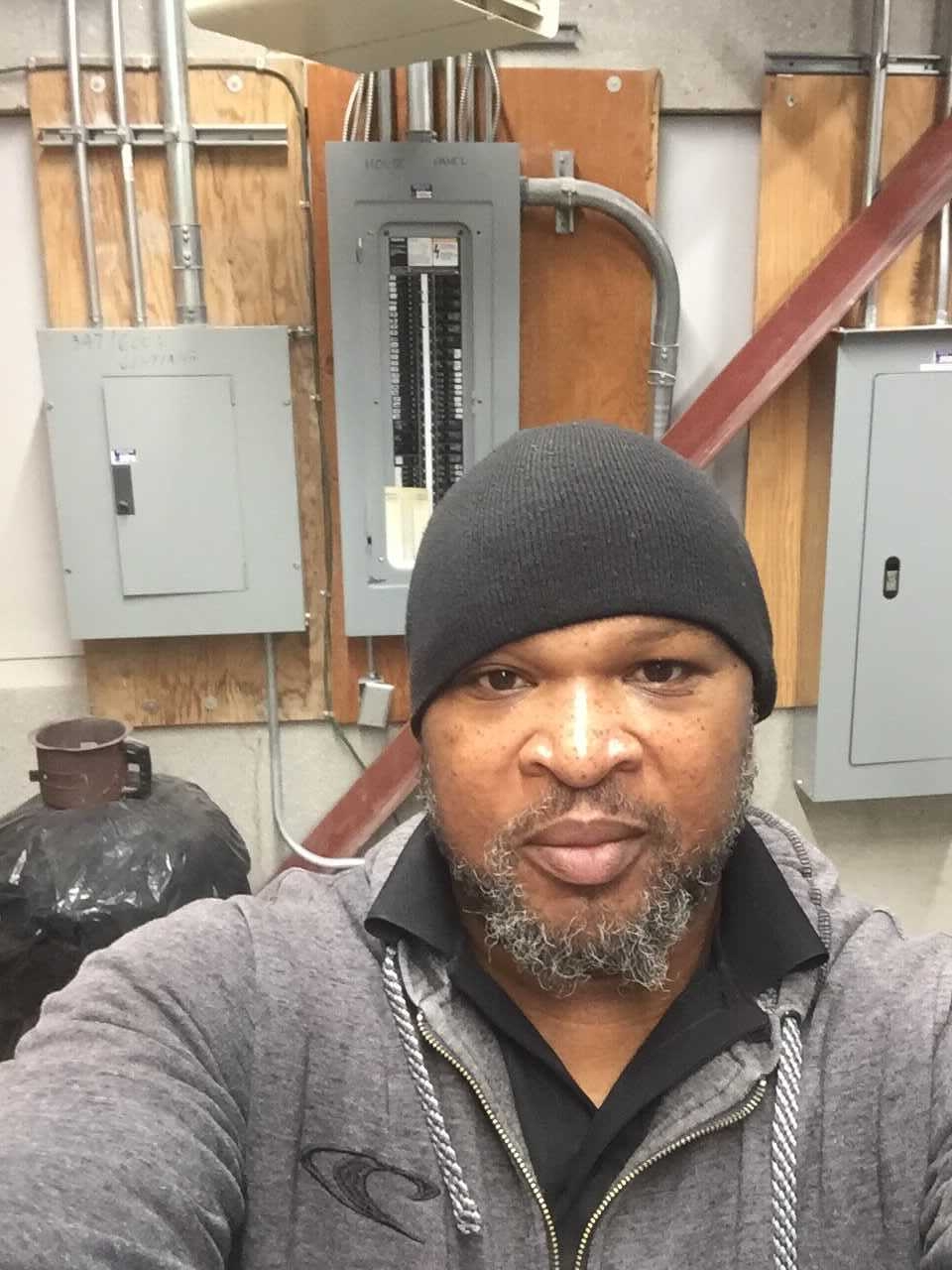 霍华德·本杰明在电气室拍的照片。霍华德穿着一件灰色的毛衣，戴着一顶黑色的针织帽。背景是保险丝盒。