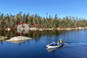 在一个阳光明媚的日子，平静的湖面上，有三个人坐在灰色的独木舟上。附近有一块岩石凸出水面，在背景中，花岗岩点缀着海岸线，绿色和黄色的树木和红色的越橘就长在后面。湖水很平静，树木在湖中倒映。