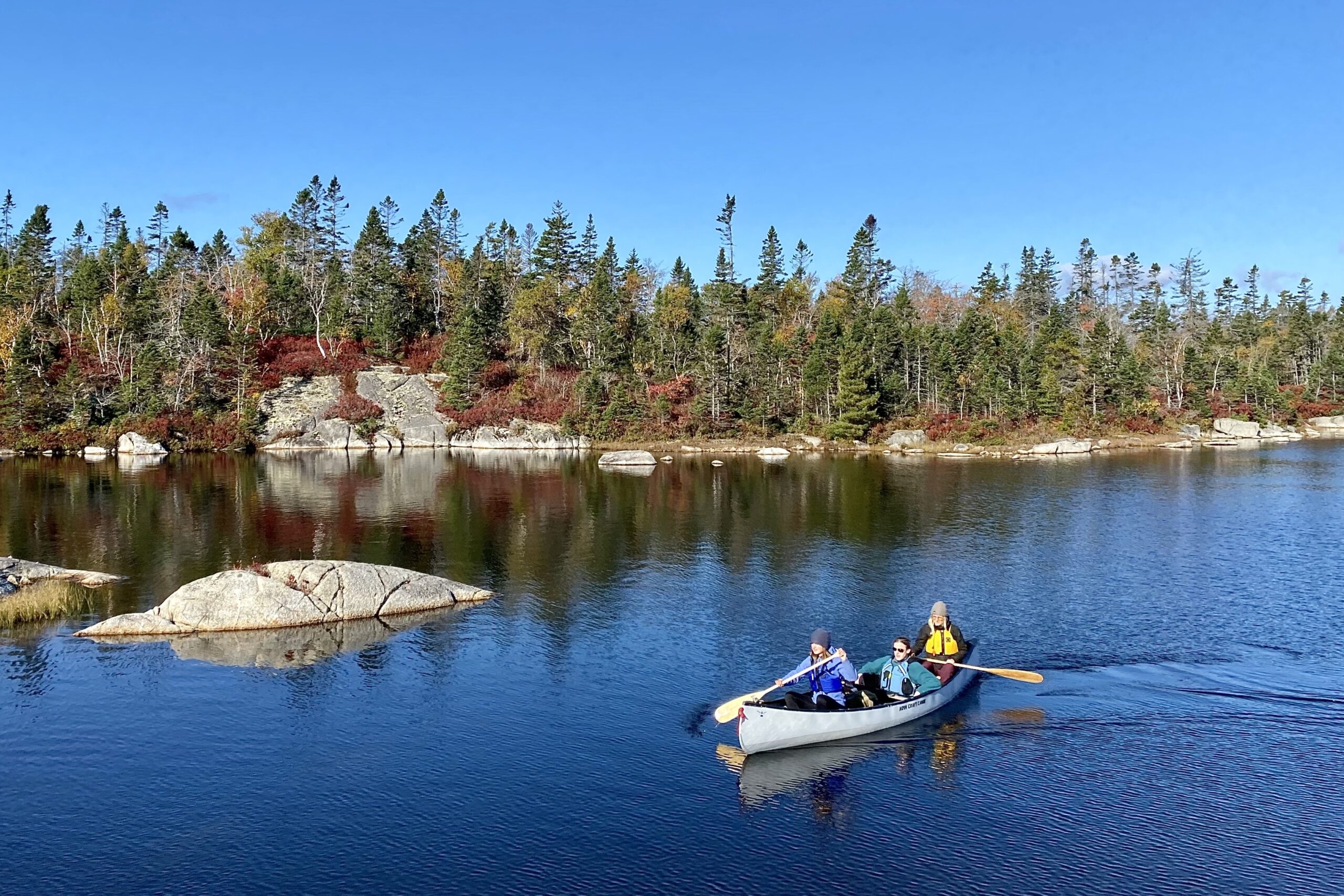 在一个阳光明媚的日子，平静的湖面上，有三个人坐在灰色的独木舟上。附近有一块岩石凸出水面，在背景中，花岗岩点缀着海岸线，绿色和黄色的树木和红色的越橘就长在后面。湖水很平静，树木在湖中倒映。