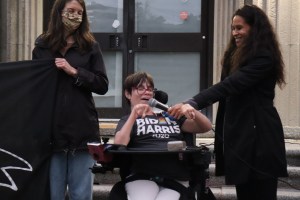 埃尔·琼斯的照片他是罗伯特·戴维特追悼会的主持人。琼斯拿着麦克风，让同样参加追悼会的维姬·莱瓦克发言。莱瓦克是当地的残疾人活动家，他使用轮椅。在莱瓦克的左边是另一个举着横幅的女人。