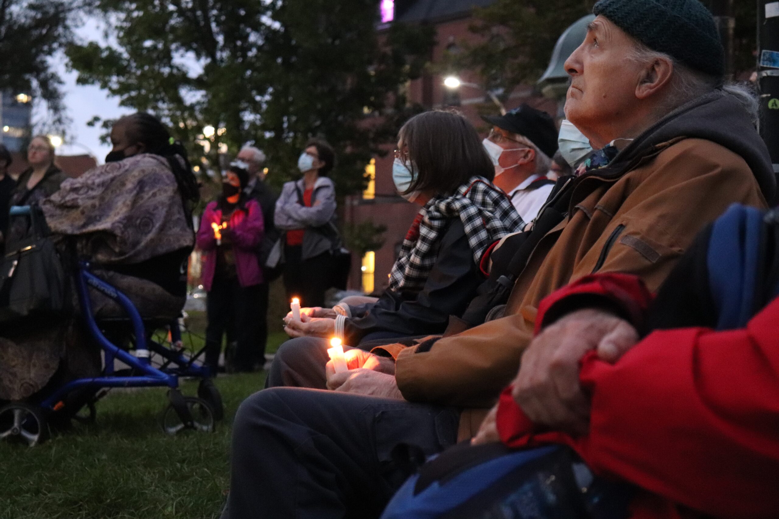 一群人聚集在Robert Devet的纪念碑前的照片，Robert Devet是新斯科舍省《倡导者》的出版商和编辑。人们或站或坐在椅子上，戴着口罩，举着蜡烛。