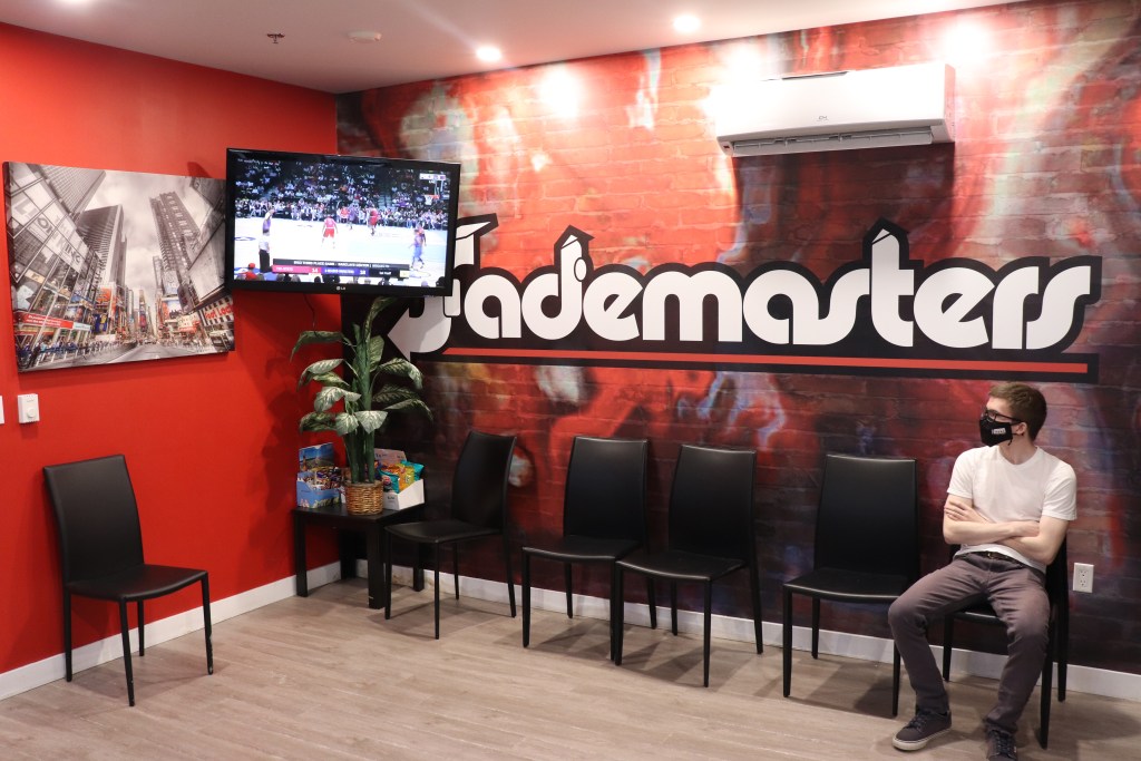 一位顾客在Fademasters理发店等待理发，理发店的logo是由业主/运营商Tremayne“Trobiz”Howe设计的。大堂被漆成橘红色，沿墙有黑色的椅子。电视安装在房间角落的墙上。一个年轻人坐在椅子上，戴着面具，穿着白色的t恤和灰色的裤子。他在看电视。