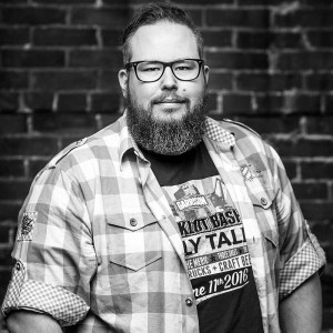 贾斯汀·辛克的黑白照片，他是哈利法克斯加里森酿造公司的营销和品牌经理。贾斯汀穿着格子衬衫，留着胡子，戴着眼镜。他站在一堵砖墙前。