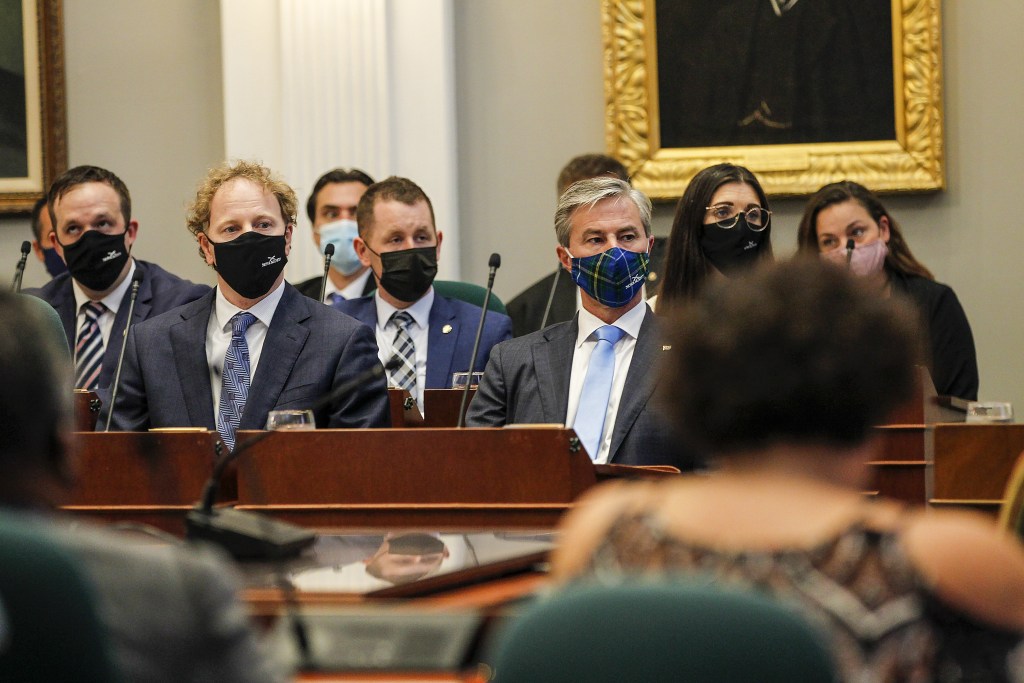 A group of people in suits and face masks sitting at wooden desks with microphones look to their left. In the background is a painting in a gold frame. The walls are beige with white trim.