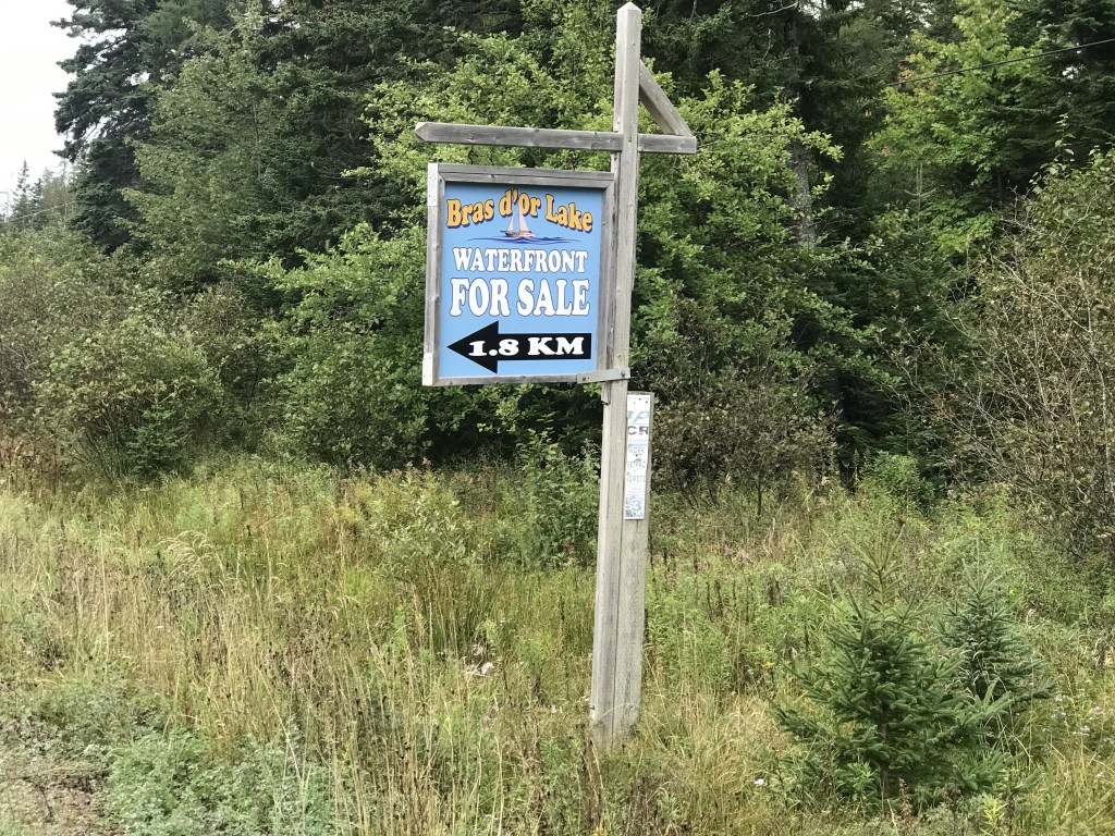 一张出售标志的照片，上面写着“Bras d’or海滨出售，1.8公里”，箭头指向左侧。该土地位于布雷顿角里士满县埃文斯岛。这个标志是在路边的一个沟里，周围有树和草。