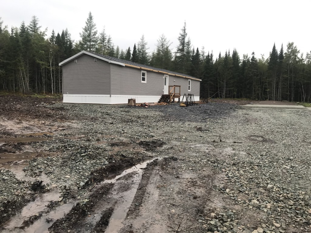 海狸小屋地产地段与预制房屋。停车场被清理干净了，有一个砾石和泥泞的区域，轮胎的痕迹贯穿其中。这栋预制房屋有一扇门、两扇窗、一个木质的前台阶和灰褐色的乙烯基壁板。在装配式房屋后面是一片树林。图片:Joan Baxter