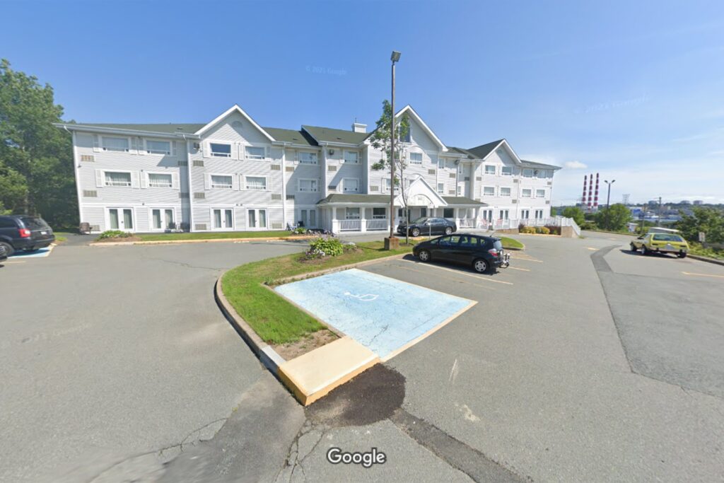 阳光明媚的一天，在停车场后面可以看到屋顶有三个山峰的白色酒店建筑。在背景中，可以看到达特茅斯的新斯科舍电厂的三个红白相间的烟囱。停车场里有几辆车，还有绿油油的林荫大道。