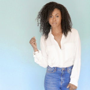 Vinessa Antoine，一名黑色妇女，带着肩长卷发，站在苍白的蓝绿色背景，穿着牛仔裤和一件白衬衫