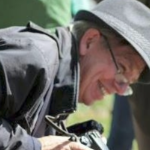 戴着灰色帽子、穿着灰色外套的罗伯特·德维特看上去衣冠楚楚，手里拿着相机微笑着。