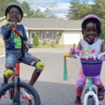 Two Black kids, aged about 6 to 8, happily pose for the camera with their bikes, wearing their bright helmets and kneepads