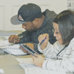 A Black man and a Black woman study with their books, papers, and a tablet at a table