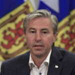 蒂姆·休斯顿（Tim Houston）穿着一件深色外套和开放的领衬衫，没有领带，在新斯科舍省一些旗帜前的讲台上讲话。