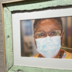 A Black woman with glasses and a blue medical mask is shown in a photo framed in a green painted frame made from reclaimed wood.