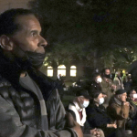 a Black man listens solemnly while a crowd sits and stands in the background at the nitghttime memorial for Robert Devet