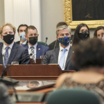 A group of newly-elected politicians, men and women, sit behind desks while wearing masks.
