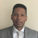 一个黑人的爆头，戴着小胡子的黑人穿着灰色的西装在米色的墙壁前。