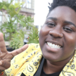 一位身穿黄黑相间夹克的黑人妇女微笑着对着镜头做和平手势。