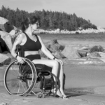 一张黑白照片，一个女人穿着黑色两件套泳衣，坐在轮椅上躺在沙滩上