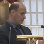 环境部长蒂姆·哈尔曼(Tim Halman)是一名秃顶的白人，身穿深色西装和深蓝色衬衫，在讲台上发言。在他的周围，是镀金相框的古老画作，红色天鹅绒窗帘，还有一把金色扶手椅，椅背上有一顶皇冠。