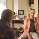 两个黑人，保罗·拜德和休伯特·戴维斯，在客厅里谈话，左边有一盏明亮的摄像机灯。