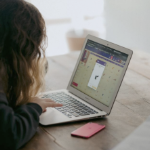 一个年轻的女孩，棕色的卷发遮住了她的脸，正在木桌旁的笔记本电脑上工作。