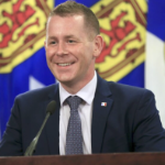 Colton LeBlanc，一个穿着海军蓝西装，白色衬衫和灰色领带的白人男子站在讲台上微笑。背景为三面新斯科舍省国旗，分别为蓝、黄、红三色。