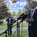 蒂姆·休斯顿在一个阳光明媚的日子里对着外面的一排麦克风讲话。背景中，一名男子正在拍照。