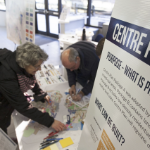 人们站在桌子前，指着一张彩色地图。他们在室内，但阳光从窗户照进来。在前景中有一个海报板，上面写着:“中心计划，目的——B包是什么?”