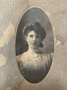 这是一幅大约100年前的女性黑白肖像。那个女人穿着一件高领白色蕾丝衬衫，头发盘了起来。这张照片是在哈利法克斯Hydrostone地区一所房子的墙上发现的。房主们正在寻找照片中女子的身份。