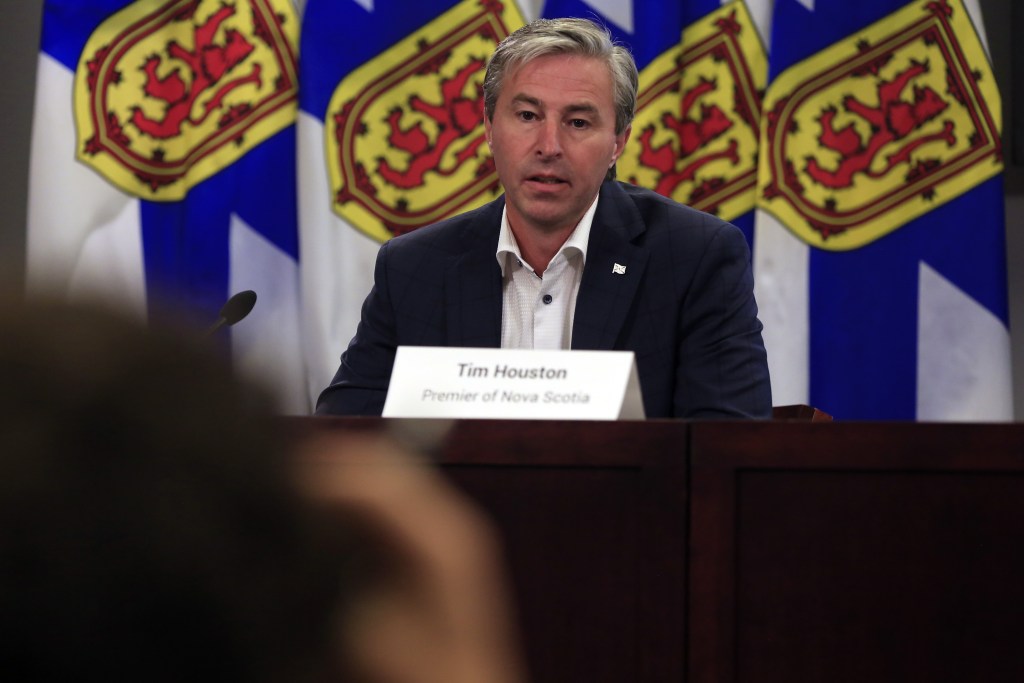 一个穿着深色西装、白衬衫、不打领带的男人在讲台上讲话。背景是四面新斯科舍省国旗，颜色分别为蓝色、黄色和红色。