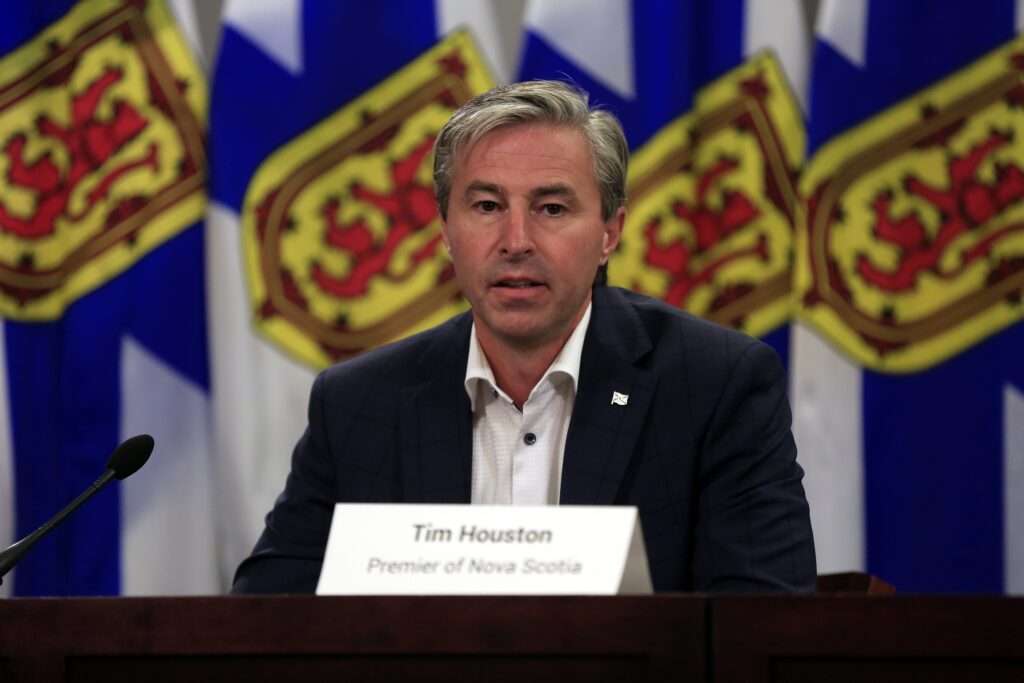 一个穿着深色西装，白衬衫和没有领带的男人在讲台上说话。背景中有四个新斯科舍省旗帜，蓝色，黄色和红色。“width=