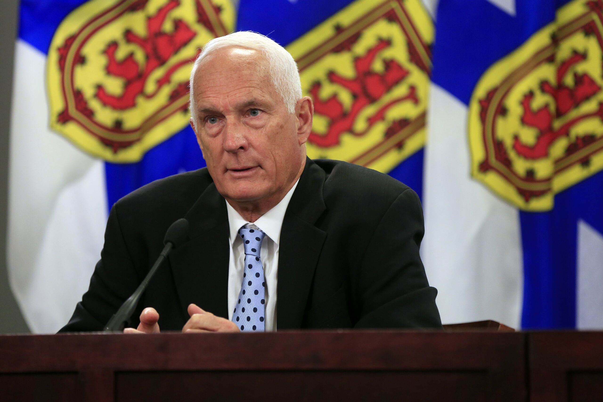 A man wearing a black suit, white shirt and blue polka dot tie speaks at a podium. In the background are three Nova Scotia flags, coloured blue, yellow and red.