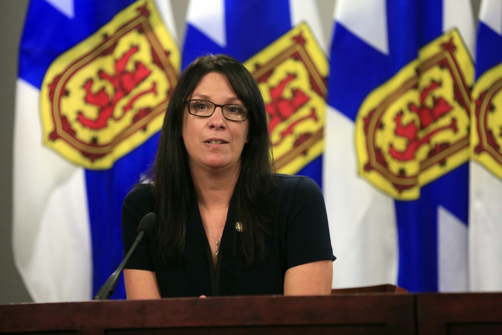 A woman wearing a dark t-shirt and glasses speaks at a podium. In the background are three Nova Scotia flags, coloured blue, yellow and red.