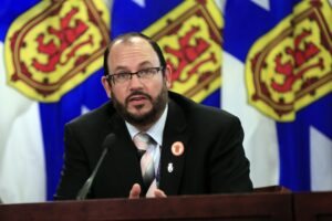 一名身穿黑色西装、条纹衬衫、粉红和黑色条纹领带、戴着眼镜的男子在讲台上对着麦克风讲话。背景是三面新斯科舍省国旗，颜色分别为蓝色、黄色和红色。