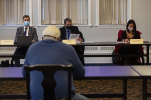 在前景中，一个男人坐在一张桌子旁，背对着镜头。他戴着一顶白色的棒球帽和一件蓝色的毛衣。背景中有三个人，两个穿西装的男人和一个戴着口罩的女人，坐在另一张桌子旁。左边的人正在看前景中的人。中间的男人在看一张纸，右边的女人在低头看桌子。