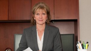 金·阿代尔(Kim Adair)坐在她的办公桌前，面前是一个带门的樱桃木书柜。她是一个白人女人，金黄色的长发及下巴，穿着灰色夹克和白色衬衫，上面有灰色的圆点。