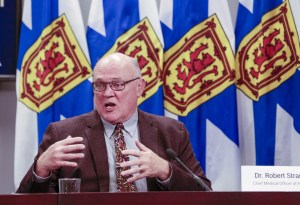 罗伯特·斯特朗博士在COVID通报会上讲话的照片。他坐在桌子旁，身后是几面蓝白相间的新斯科舍国旗。斯特朗穿着棕色夹克，米色衬衫，打着花纹领带。