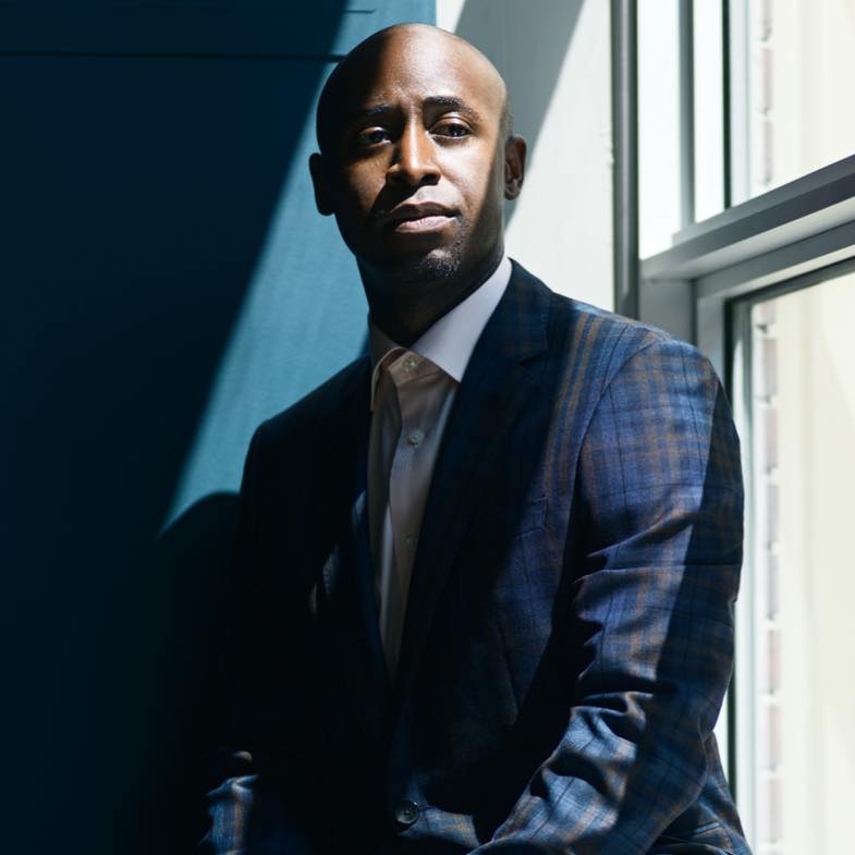 这是一张查德·威廉姆斯医生坐在窗边的照片，有几束光线照进来。威廉斯博士穿着一套蓝色格子呢西装和浅蓝色衬衫。