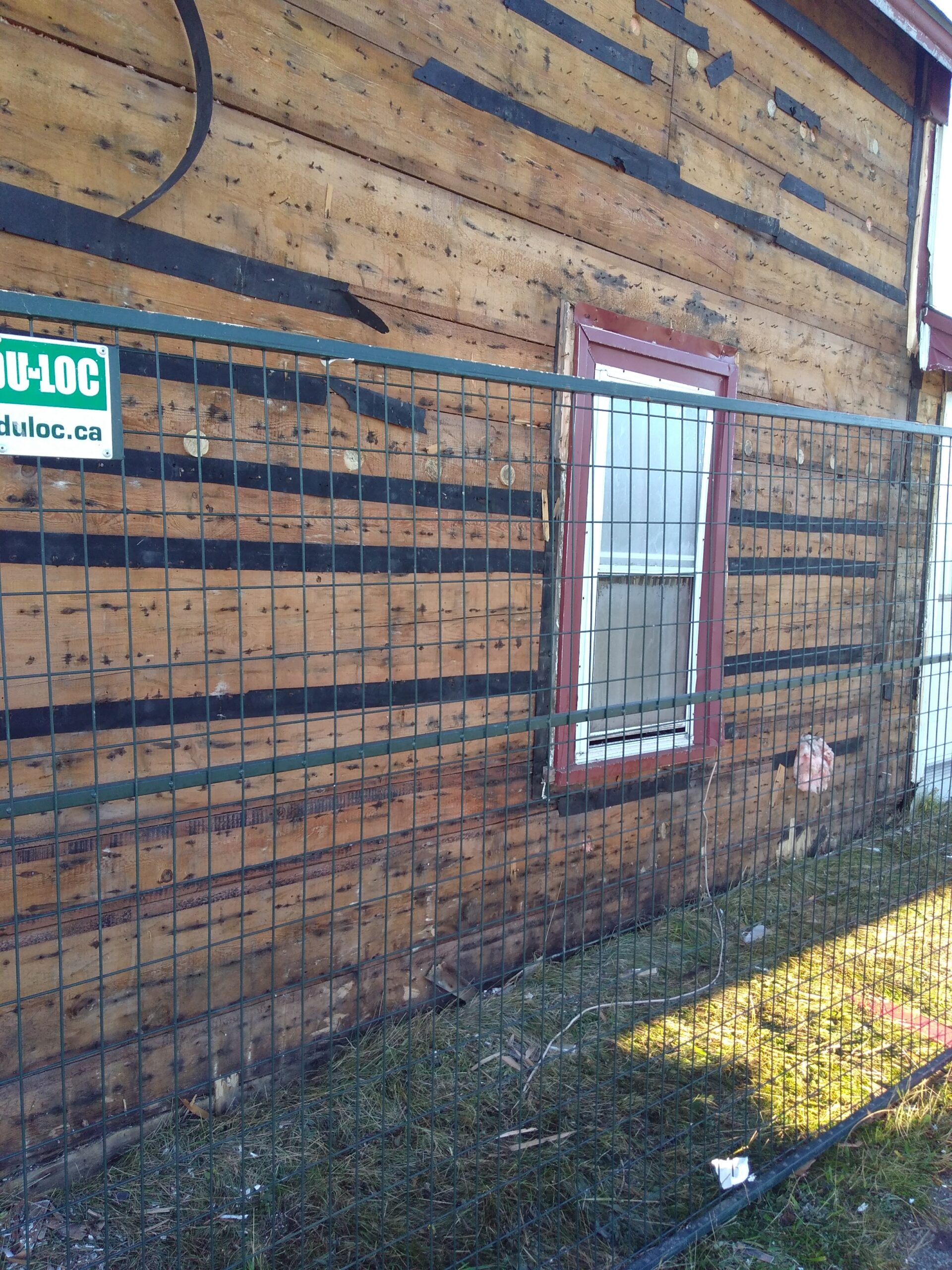 近景的房子的一面与墙板拆除和长宽旧板可见。房子在一个临时的金属栅栏后面。