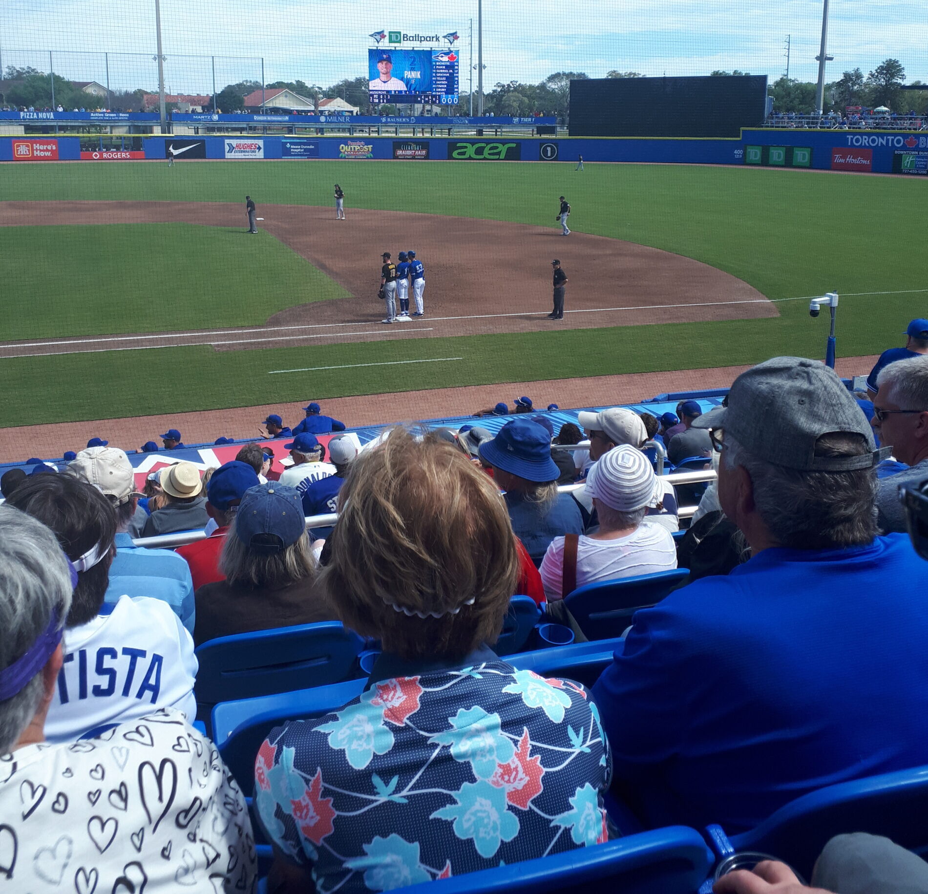 这张照片是在一个小棒球场的一垒边的座位上拍摄的。这是一个晴朗的晴天。人们坐在看台上，可以看到何塞·包蒂斯塔的球衣。一垒上有一个跑垒者。