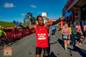 Dennis Mbelenzi首先在哈利法克斯的ScotiaBank中心外的2021年Bluenose马拉松，在两小时半小时的一分钟害羞，前方15分钟前，第2位赛道。Mbelenzi伴随着他的武器，旨在庆祝他的胜利。马拉松路线在Citadel Hill下面的Brunswick街上。他身后还有其他跑步者和街道上的人群。