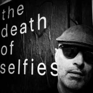 安德鲁·墨菲的黑白自拍照。他是一个白人，戴着深色太阳镜，戴着报童风格的尖顶帽，在他身后的黑色墙上用白色字体写着“自拍的死亡”。