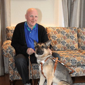 查理和他以前的导盲犬桃子坐在沙发上。查理是一个上了年纪的白人，笑到一半，桃子是一只德国牧羊犬，沙发上是花。