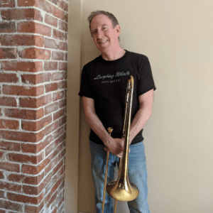 丹尼·马丁(Danny Martin)是一个微笑着的白人，穿着黑色t恤和牛仔裤，靠在墙上，手里拿着他心爱的长号。