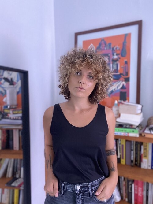 一个穿着黑色背心的女人，一头卷发，站在客厅里，身后是一幅画和几本书。