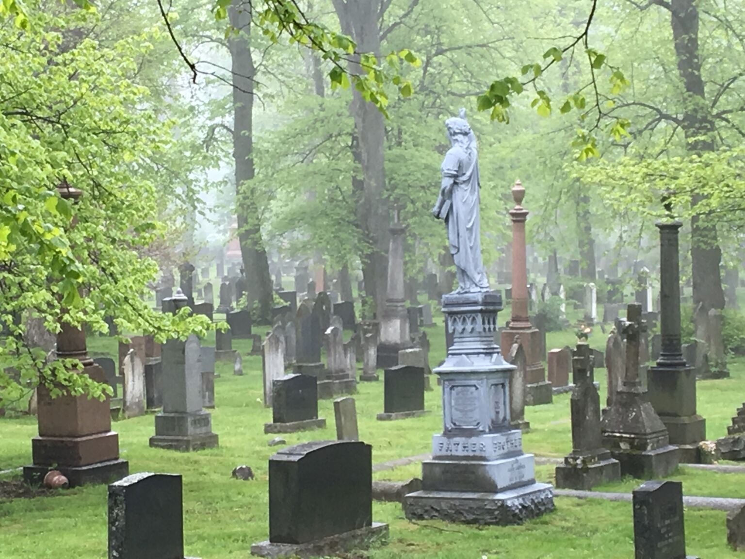 公墓里有几块低矮的墓碑和一座更高的锌制白色纪念碑，上面有一个人站在基座上。背景中有树。