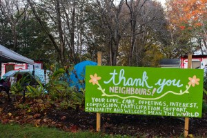 在海法克斯的米格尔公园外，写着“谢谢你，邻居!”的绿色标志。标志后面立着帐篷，部分被公园花园遮挡。